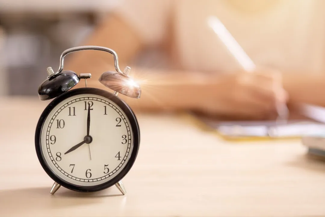 Close-up of a classic black alarm clock on a study desk with soft sunlight. In the blurred background, a student is writing on paper, symbolizing timed practice or focused study.